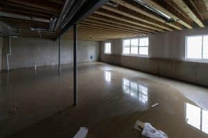 A flooded basement with standing water covering the floor, exposed ceiling joists and pipes, and windows reflecting in the water.