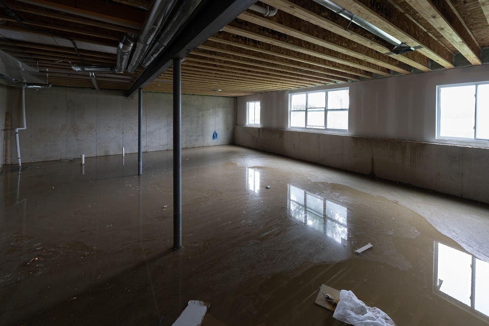 A flooded basement with standing water covering the floor, exposed ceiling joists and pipes, and windows reflecting in the water.